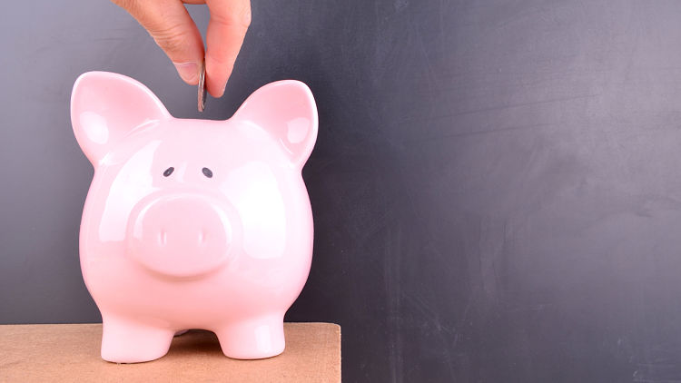 A coin is deposited into a pink piggybank with a blank blackboard in the background.