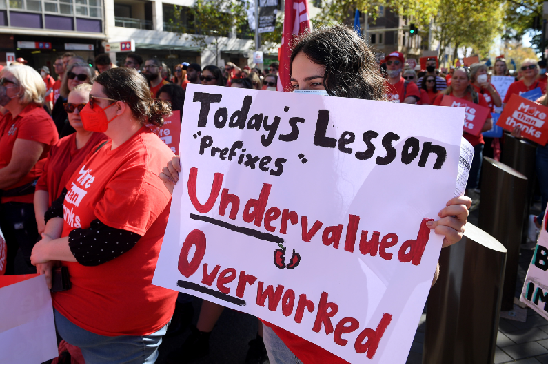 teacher with a sign saying 'undervalued', 'overworked'